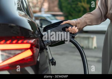 Le connecteur de charge de véhicule électrique est inséré dans le port de charge d'une voiture noire élégante. La scène se déroule à une station de recharge publique avec des véhicules flous en arrière-plan. Banque D'Images