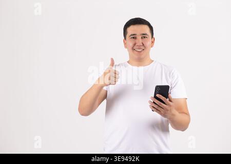Homme heureux en t-shirt blanc tenant le téléphone et montrant le pouce vers le haut Banque D'Images