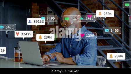 Homme chauve à barbe blanche travaillant dans la chemise en denim tapant sur ordinateur portable dans le loft, le verre et les notifications Banque D'Images