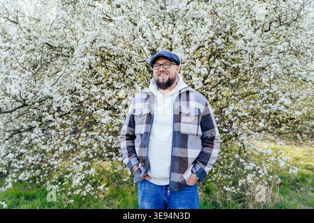 Homme caucasien barbu souriant en surpoids dans des lunettes debout près de l'arbre en fleurs Banque D'Images