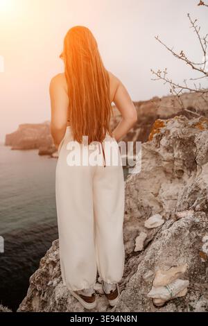 Femme falaise océan jeune femme avec de longs cheveux se tient sur une falaise rocheuse surplombant l'océan calme au coucher du soleil Banque D'Images