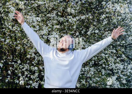 Homme barbu heureux portant des écouteurs avec les bras levés près de l'arbre en fleurs Banque D'Images