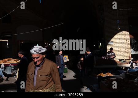 Un homme portant un turban blanc et une veste marron marche à travers un marché faiblement éclairé sous un bâtiment en briques. La lumière du soleil coule à travers le toit, coulant l'alose Banque D'Images