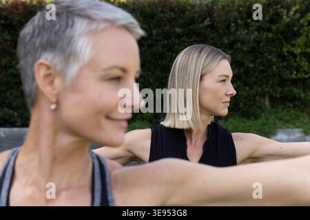 Amies féminines s'étirant sur la pelouse herbeuse dans la cour arrière portant des débardeurs près de la haie taillée et du mur. Extérieur, fitness, yoga, stretch, duo, nature, bien-être Banque D'Images