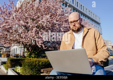 Homme réfléchi utilisant un ordinateur portable dans le parc de la ville pendant la journée de printemps sous l'arbre en fleurs Banque D'Images