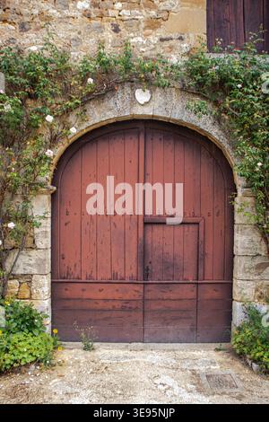 Une charmante vue sur une ancienne porte en bois voûtée, riche en histoire, ornée de roses grimpantes et de lierre, située dans un mur de pierre dans le village médiéval de Banque D'Images