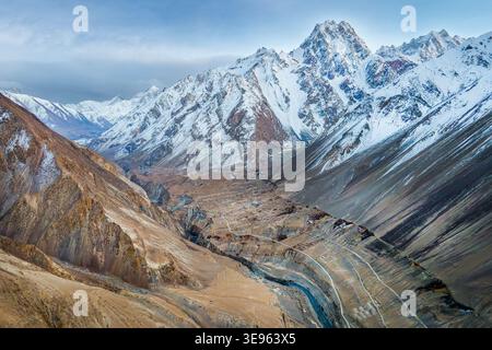 Vue aérienne des sommets escarpés et enneigés qui s'élèvent au-dessus du fond de la vallée aride, où un mince ruban de rivière sculpte à travers le paysage, Hunza Nag Banque D'Images
