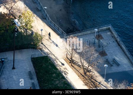 Manhattan, États-Unis - 1er décembre 2025 : vue d'un personnage solitaire se promenant le long du front de mer de l'East River, coupé en deux par la lumière du soleil et les ombres, Banque D'Images