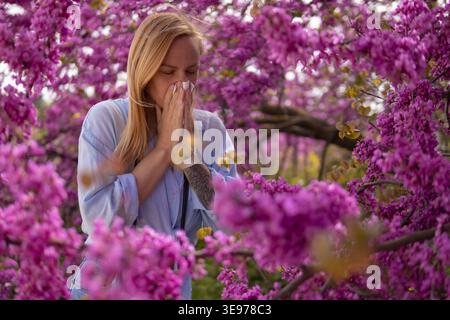 Jeune femme debout dans un parc. Souffrant d'allergies saisonnières sévères. Éternuant et soufflant son nez avec un tissu blanc entouré par l'abondant Banque D'Images