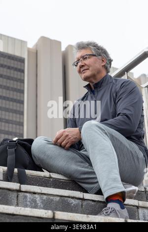Homme senior assis sur des marches en béton dans la place urbaine portant des lunettes, sac à dos reposant sur des marches Banque D'Images