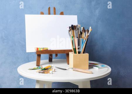 Pinceaux, couteau, peintures et chevalet avec toile sur table près du mur bleu Banque D'Images