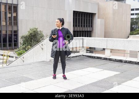 Femme mature debout sur le toit au rail de béton, tenant tapis vert, téléphone, manteau matelassé, casque Banque D'Images