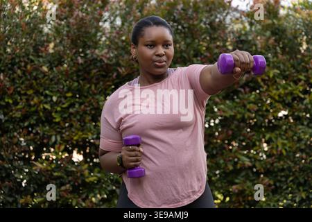 Femme afro-américaine exerçant avec des haltères violets dans le jardin en tee-shirt rose et leggings foncés Banque D'Images
