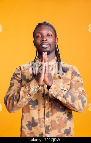 Portrait d'un homme paisible priant dieu demandant pardon tout en se tenant dans un studio avec fond jaune. Personne religieuse afro-américaine tenant les paumes ensemble dans Pray. Concept de religion Banque D'Images