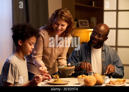 Famille multiethnique partageant le dîner à la maison, servant de la nourriture et mangeant ensemble. Représentation parentale, liens familiaux, routine des repas, publicité sur le style de vie, plan moyen Banque D'Images
