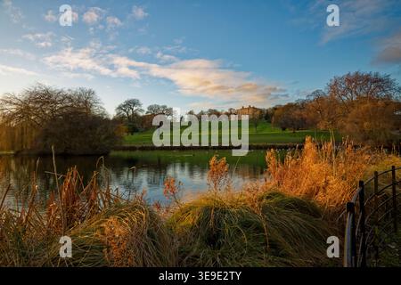 Un paysage de parc avec un lac et une grande maison de campagne sur une crête illuminée par la lumière douce du soir, Cusworth Park, Doncaster, South Yorkshire. Banque D'Images