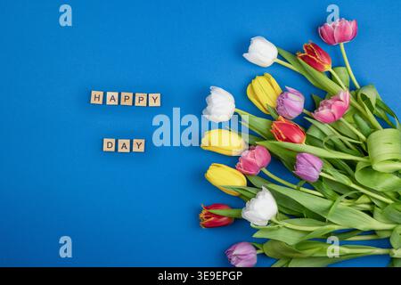 Fond de lettres en bois de texte Happy Day. Bouquet de fleurs de tulipes sur couleur bleue. Mots de la vue de dessus. Bourgeons multicolores floraux Banque D'Images