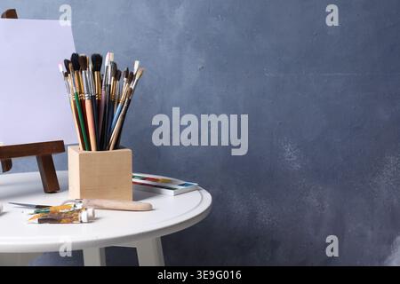 Pinceaux, couteau, peintures et chevalet avec toile sur table près du mur bleu. Espace pour le texte Banque D'Images