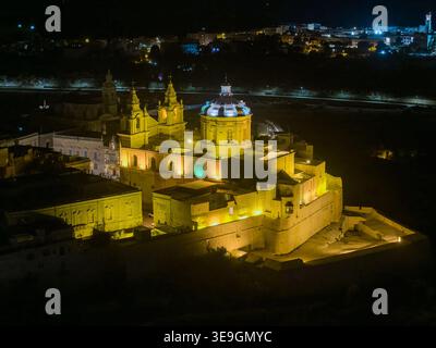 Vue nocturne sur la cathédrale de Paul et les murs fortifiés de Mdina illuminés par une lumière chaude, surplombant le paysage maltais sombre. Banque D'Images