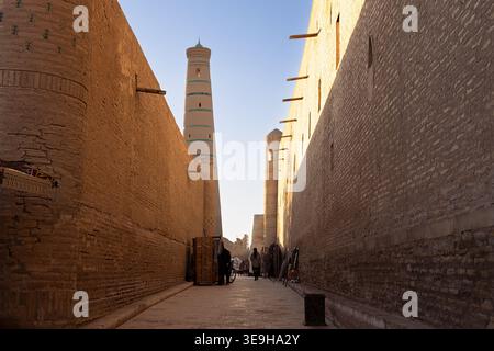 Ruelle étroite dans la vieille ville d'Ichan Kala de Khiva avec minaret s'élevant au-dessus des murs de briques. La lumière chaude de l'heure dorée illumine l'ancien paysage de rue, idéal Banque D'Images