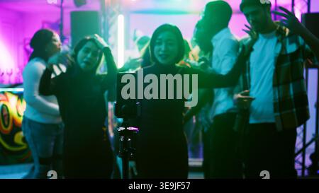 Femme filmant vlog pour les médias sociaux, en utilisant smartphone à la boîte de nuit sur la piste de danse. Jeune adulte enregistrant une vidéo amusante d'amis dansant et faisant la fête ensemble, divertissement. Prise de vue à main levée. Banque D'Images