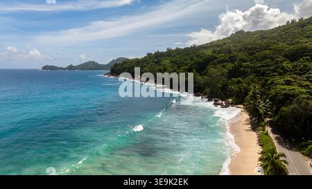 Les vagues se baignent doucement sur un rivage sablonneux entouré de collines verdoyantes et d'eaux bleues claires, sous un ciel lumineux. Seychelles, Mahé. Anse Takamaka Beach. Banque D'Images