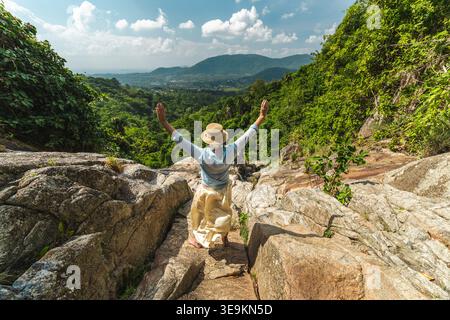 Jeune femme avec les bras levés debout sur le chemin rocheux surplombant la vallée tropicale et les collines lointaines Banque D'Images