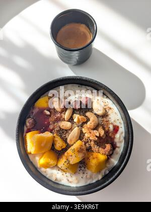 Bol de gruau de petit déjeuner sain avec mangue, noix mélangées, graines de chia, confiture et tasse de café espresso noir dans la lumière du soleil du matin. Banque D'Images