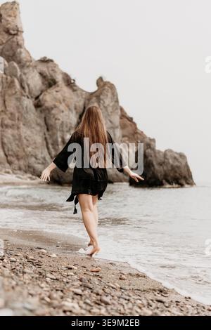 Femme plage océan marchant le long du rivage de galets avec les bras tendus vers une côte rocheuse sous un ciel brumeux Banque D'Images