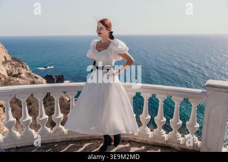 Femme robe blanche océan debout sur un élégant balcon donnant sur l'eau turquoise et les falaises rocheuses pendant des vacances méditerranéennes Banque D'Images