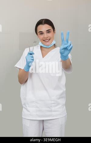 Un dentiste se tient dans un cadre neutre, souriant et tenant des instruments dentaires. Le professionnel porte des gants et un masque, transportant la préparation et Banque D'Images