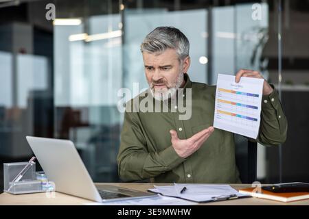 Homme d'âge moyen frustré montrant un document de mesures financières pendant un appel vidéo, discutant de problèmes commerciaux et d'analyses depuis son bureau moderne Banque D'Images
