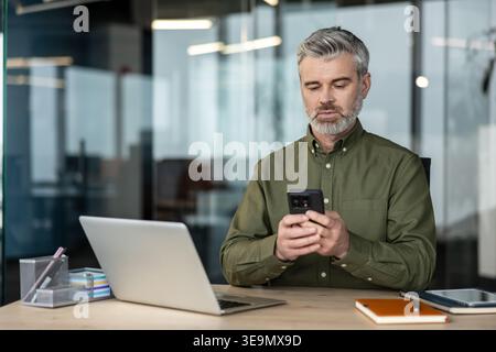 Homme d'affaires mature concentré utilisant un téléphone portable pour la communication et le travail tout en étant assis à un bureau en bois clair avec un ordinateur portable dans un cadre de bureau contemporain Banque D'Images
