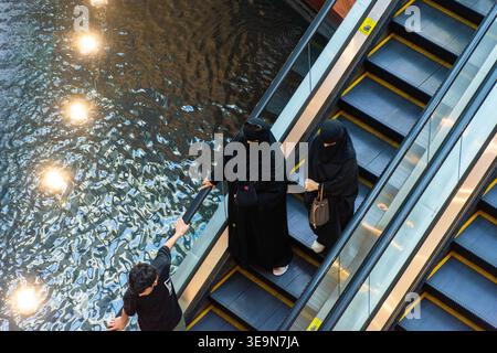 Vue en angle élevé de deux femmes musulmanes portant des abaya et niqab noires traditionnelles montant un escalier roulant descendant à côté d'un plan d'eau intérieur dans un centre commercial. Banque D'Images