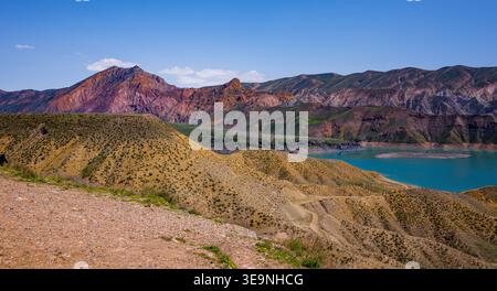 Les montagnes escarpées et brun rougeâtre surplombent le lac turquoise. Les collines roulent au premier plan, recouvertes d'herbes sèches. Ciel bleu clair, quelques nuages blancs visibles. Banque D'Images
