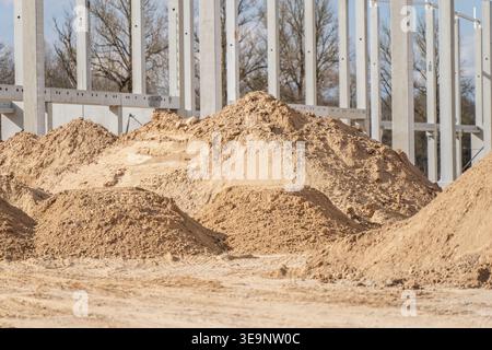De grands tas de sable et d'agrégats étagés sur le site de construction d'entrepôt à côté de la grille à colonnes en béton pour les travaux de terrassement, le nivellement et l'utilisation de l'approvisionnement en matériaux Banque D'Images