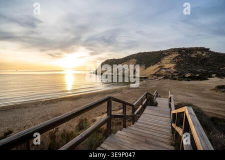 Une promenade pittoresque en bois mène l'œil vers Playa de los Cocedores au lever du soleil. Le soleil se lève sur la mer calme, illuminant les falaises dorées et Banque D'Images