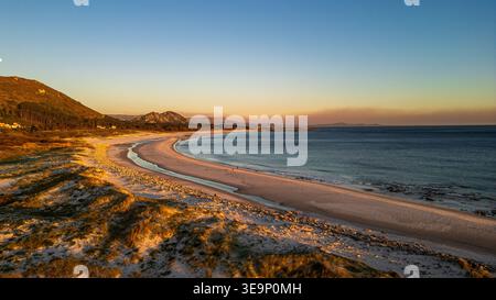 Une vue aérienne spectaculaire capture l'heure dorée sur la plage de Lariño. La lumière chaude du coucher de soleil illumine les dunes et le sable blanc, tandis que l'océan Atlantique g Banque D'Images