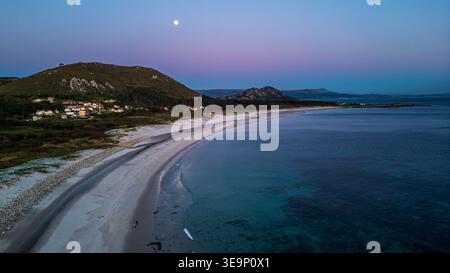 Une vue aérienne spectaculaire capture la plage de Lariño sous la lumière de la pleine lune. Le ciel crépusculaire violet pend au-dessus des montagnes et du village, tandis que th Banque D'Images
