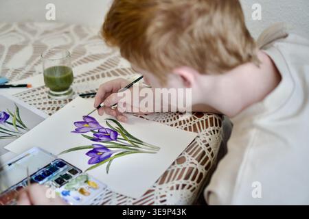 Une photo de haute qualité au-dessus de l'épaule d'un jeune homme aux cheveux clairs peignant soigneusement une illustration botanique de crocus violets. La scène montre l'artiste à l'aide d'un pinceau et d'une palette d'aquarelles sur une surface texturée. Parfait pour le matériel éducatif, les promotions des écoles d'art et le contenu de style de vie sur les loisirs et la créativité. artiste peignant des fleurs de crocus violettes avec des aquarelles. Processus créatif et concept d'éducation artistique. Photo de haute qualité Banque D'Images