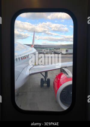 Vue d'une fenêtre d'avion montrant le moteur, l'aile et le terminal de l'aéroport sous un ciel nuageux. Banque D'Images