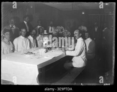Cette scène de l'expédition Flint Island de 1907 montre un dîner de Noël américain à l'intérieur d'une grande tente en toile. Une longue table est sertie de draps blancs, d'assiettes de fruits, de bouquets et de verres vides. Sept individus sont assis autour de la table, avec une femme à l'avant gauche. Au moins neuf autres hommes se tiennent autour de la table, tous habillés formellement. Un drapeau américain est visible en arrière-plan. Banque D'Images