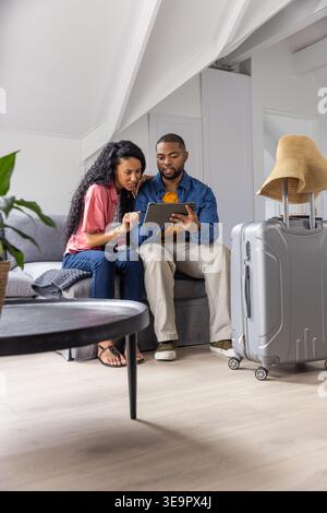 Couple diversifié assis sur le canapé gris dans le loft, étudiant tablette planifier voyage près de la valise avec chapeau Banque D'Images