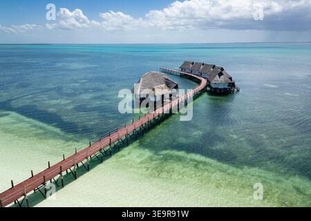 Vue aérienne de bungalows de luxe sur l'eau avec des toits de chaume et une jetée en bois entourée par l'océan émeraude et la plage de sable blanc à Zanzibar. Banque D'Images