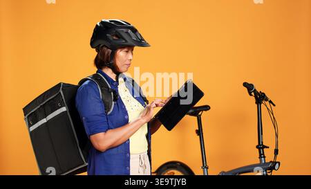 Pilote de vélo de livraison professionnel tapant les détails de la commande du client sur tablette. Courrier BIPOC portant casque de protection et sac à repas thermique vérifiant l'application en ligne, isolé sur fond de studio Banque D'Images