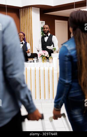 Positif amical et amical le personnel de la réception accueille les clients avec le sourire dans l'hôtel. Vue arrière d'un couple noir tirant des valises allant à la réception pour confirmer la réservation Banque D'Images