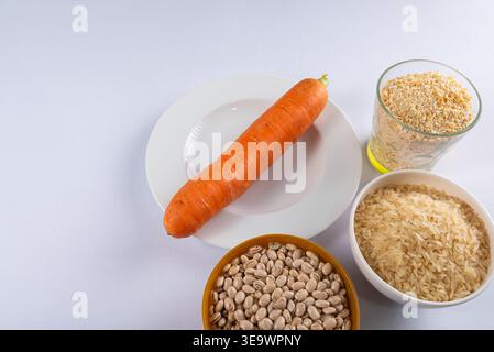 Le tir aérien présente des carottes sur assiette, divers grains et légumineuses dans des bols et du verre. Arrière-plan blanc propre. Suggère une alimentation saine, ingrédient brut Banque D'Images
