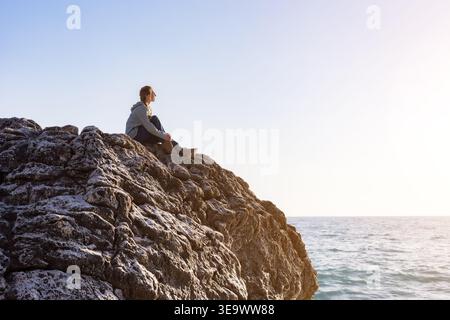 Femme solo assise sur une falaise rocheuse au bord de la mer, regardant l'horizon. Banque D'Images