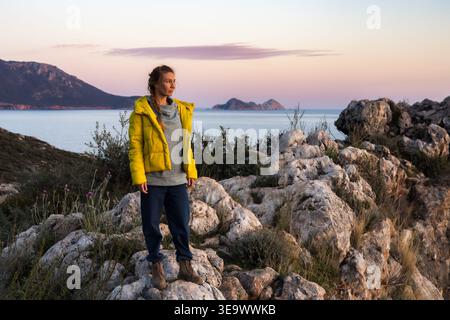 Jeune femme debout sur les rochers côtiers au coucher du soleil. Banque D'Images
