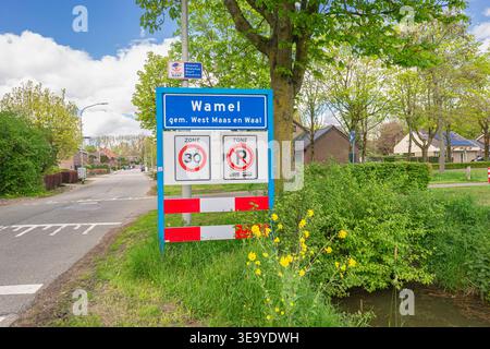 Panneau de village de Wamel montrant la zone de 30 km/h, restriction de stationnement à côté d'une rue calme, avec des fleurs de printemps jaunes et des maisons résidentielles dans un village hollandais. Banque D'Images